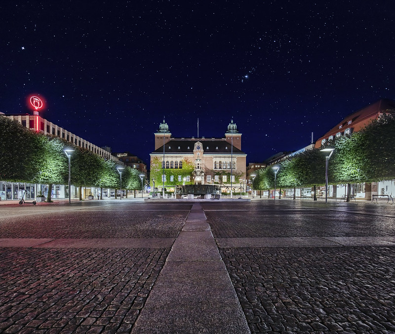 Foto på Stora torget i Borås. Fotot är tagen på kvällen, i bakgrunden syns en röd neonskylt för en biograf, torget och rådhuset är belyst av lampor.