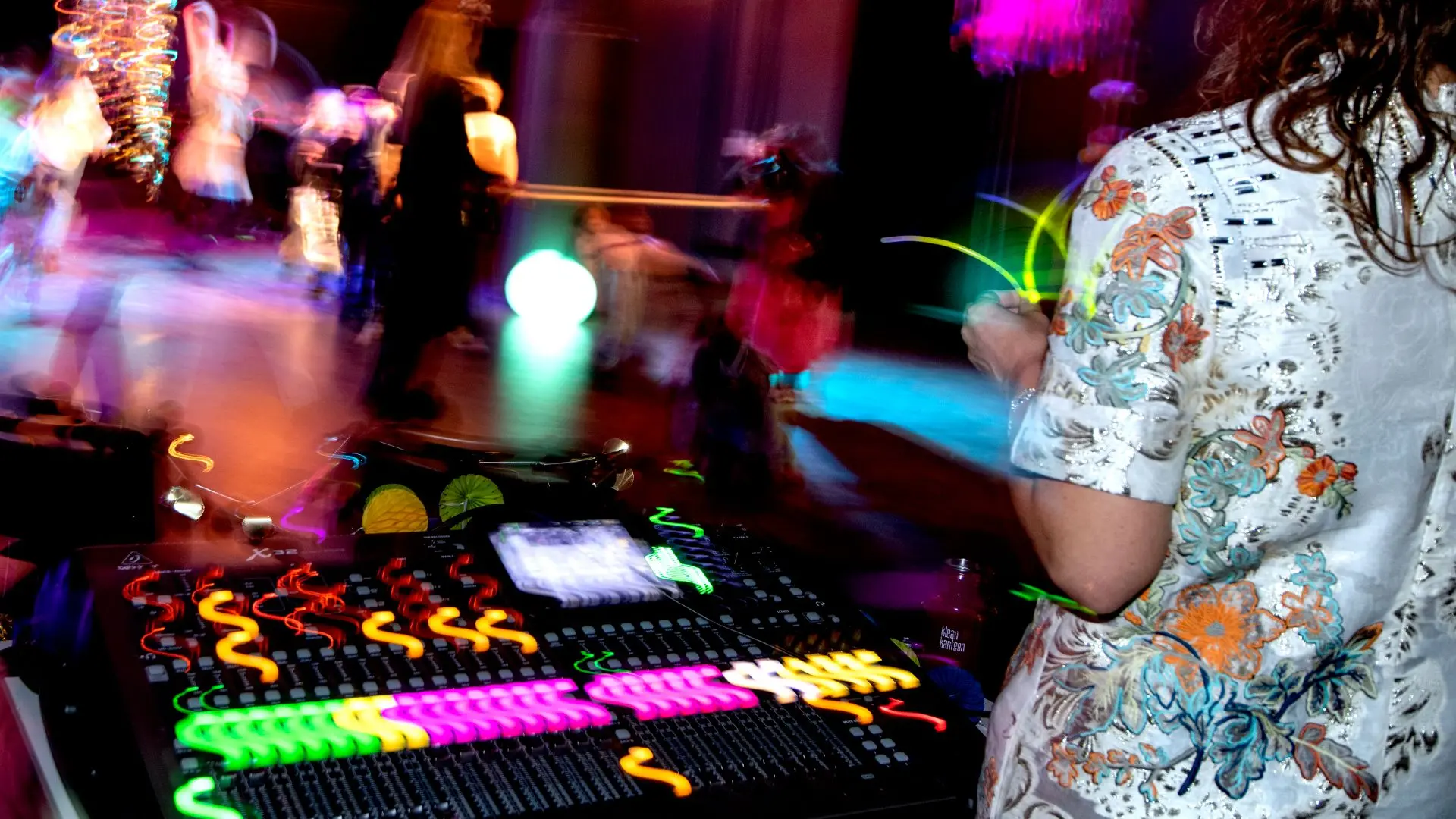 Foto på ett disco. En Dj står vid ett mixerbord, rummet är dekorerat med färgglada lampor.