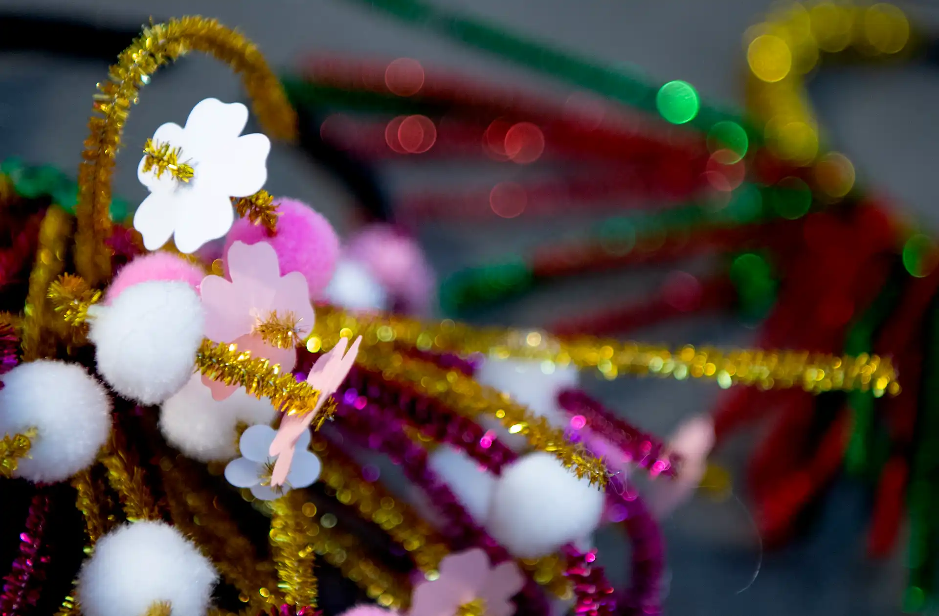 Foto på glittriga piprensare i olika färger. På piprensarna sitter vita pompom-bollar och blommor i papp. 